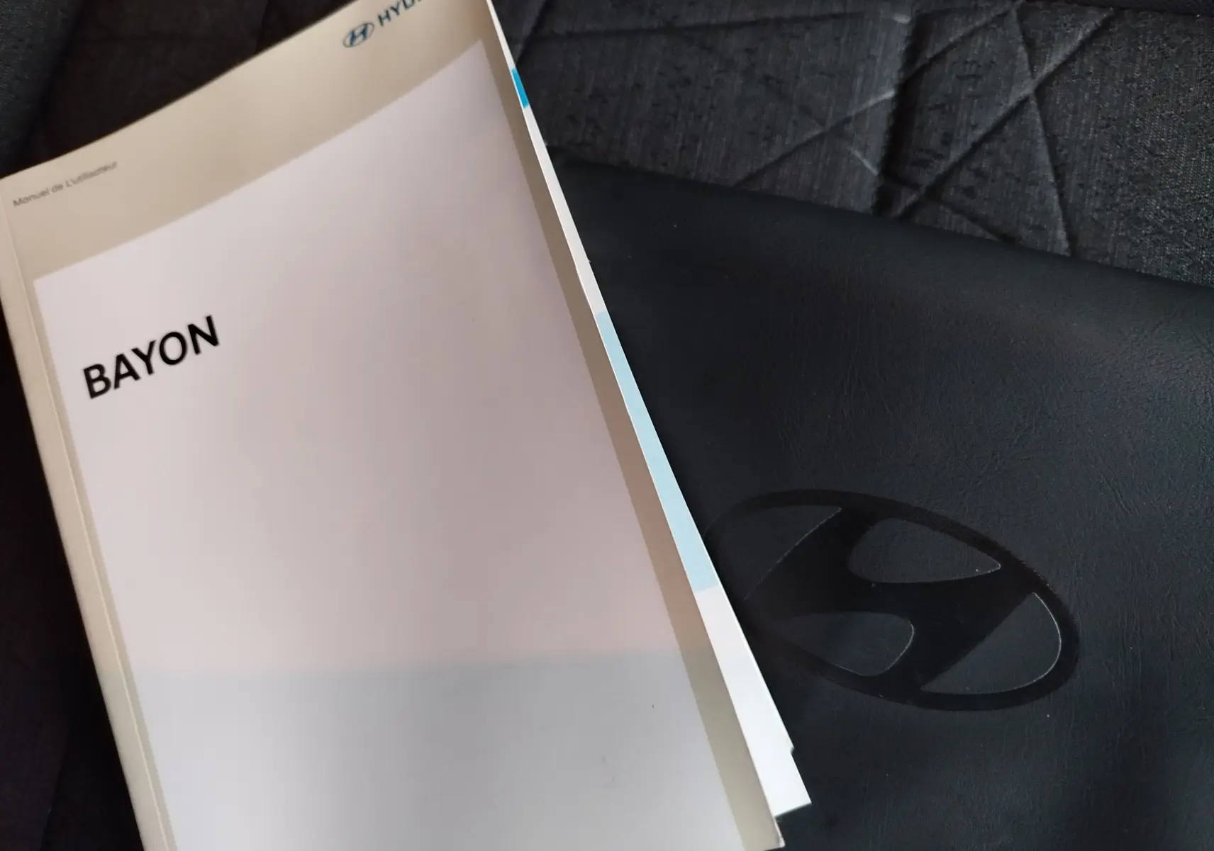 Manuel d'utilisation Hyundai Bayon posé sur un siège tissu gris avec housse en cuir noir embossée du logo Hyundai.