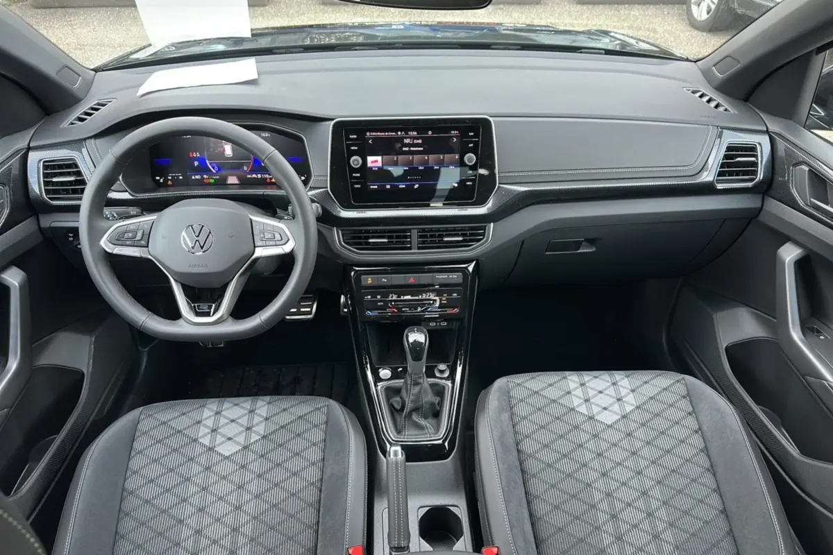 Vue intérieure avant du Volkswagen T-Cross TSI 116 DSG7 R-Line 2025, avec tableau de bord numérique et sellerie tissu grise à motifs.