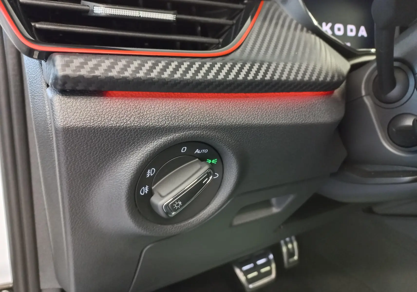 Gros plan sur le bouton de commande des phares avec finition carbone et éclairage d'ambiance rouge dans un Skoda Kamiq Blanc Lune.