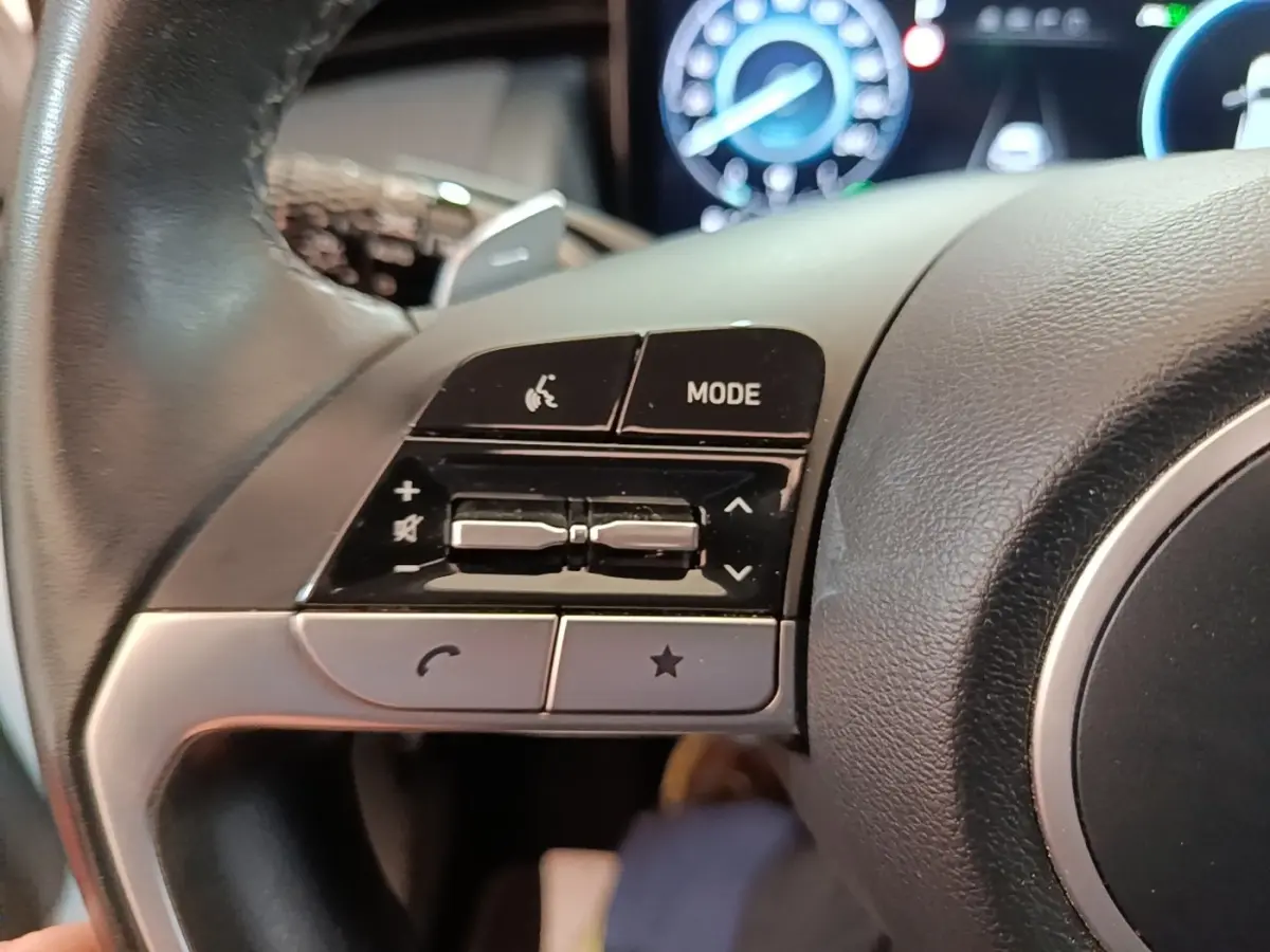 Gros plan sur les commandes gauche du volant cuir noir du Hyundai Tucson blanc hybride 2022 avec tableau de bord numérique.