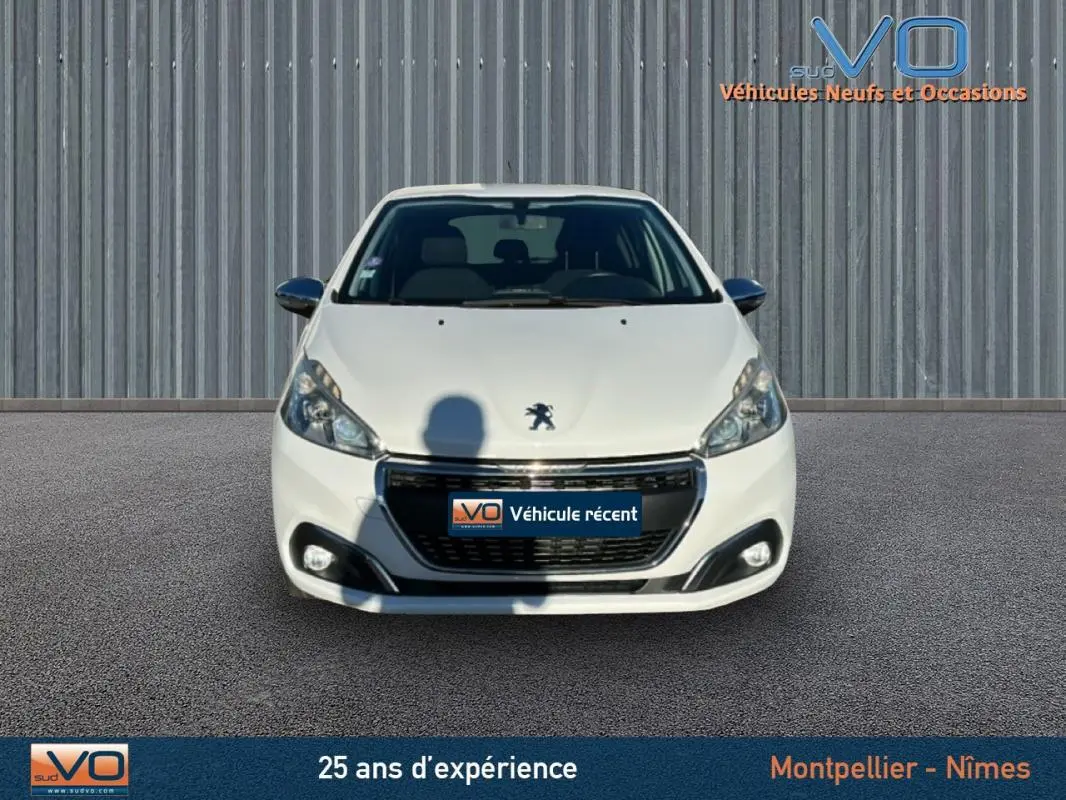 Vue avant d'une Peugeot 208 blanche de 2015, avec phares allumés et calandre noire brillante sur fond neutre.