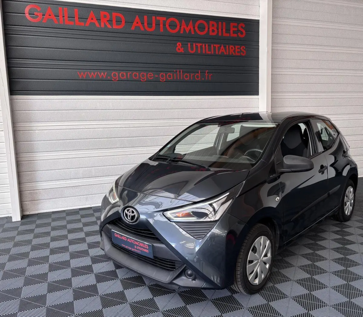 Toyota Aygo 1.0 VVT-i x-black gris foncé vue 3/4 avant droit dans un garage avec sol à damier.