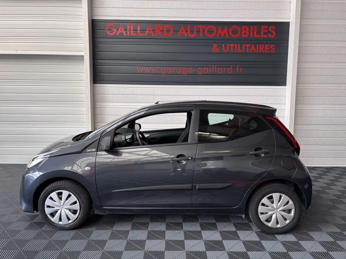 Profil côté gauche d'une Toyota Aygo 1.0 VVT-i x-black gris foncé, avec jantes standard et intérieur visible.