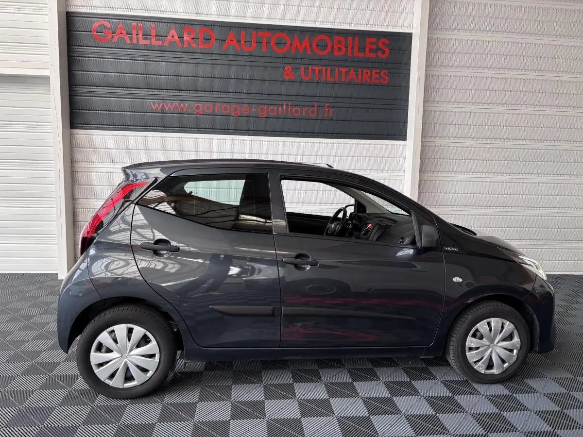 Profil côté gauche d'une Toyota Aygo 1.0 VVT-i x-black gris foncé, en intérieur sur sol damier, avec jantes argentées.