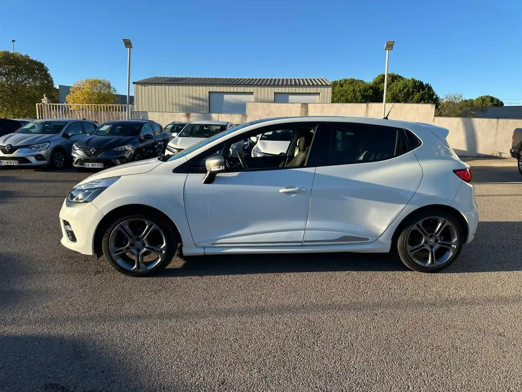 Profil côté gauche d'une Renault Clio IV blanche avec jantes alliage 17 pouces sur fond de parking ensoleillé.