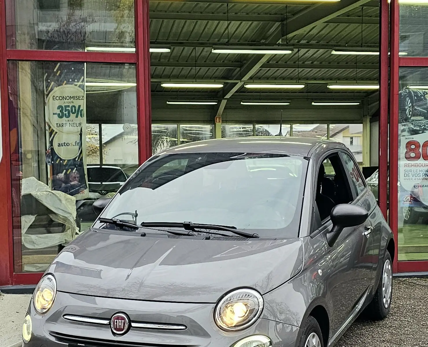 Vue 3/4 avant d'une FIAT 500 gris métallisé avec phares allumés devant un garage AD Expert.