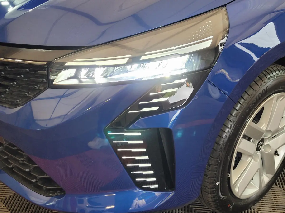Gros plan sur le phare avant droit allumé d'une Renault Clio 2025 bleu avec jante argentée et détails LED distinctifs.