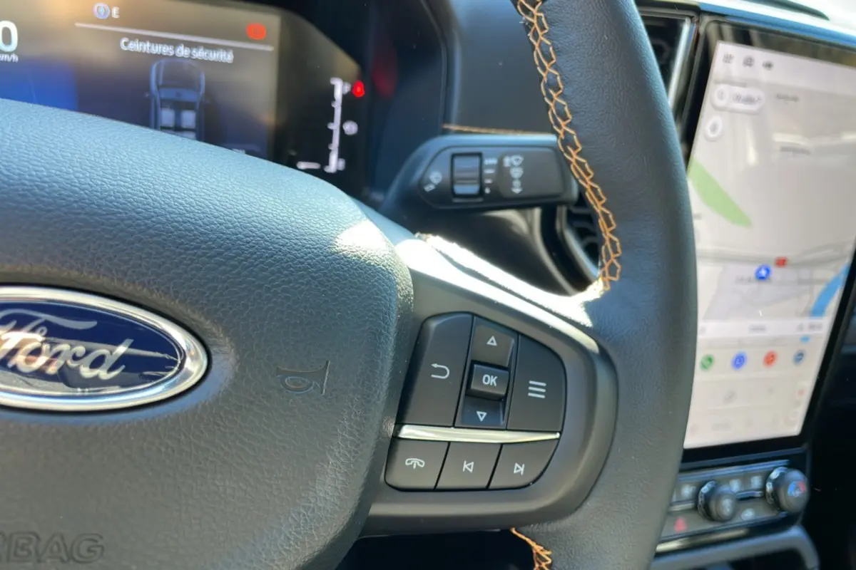 Gros plan sur le volant noir avec surpiqûres orange du Ford Ranger 2025, affichage numérique et écran tactile visible.