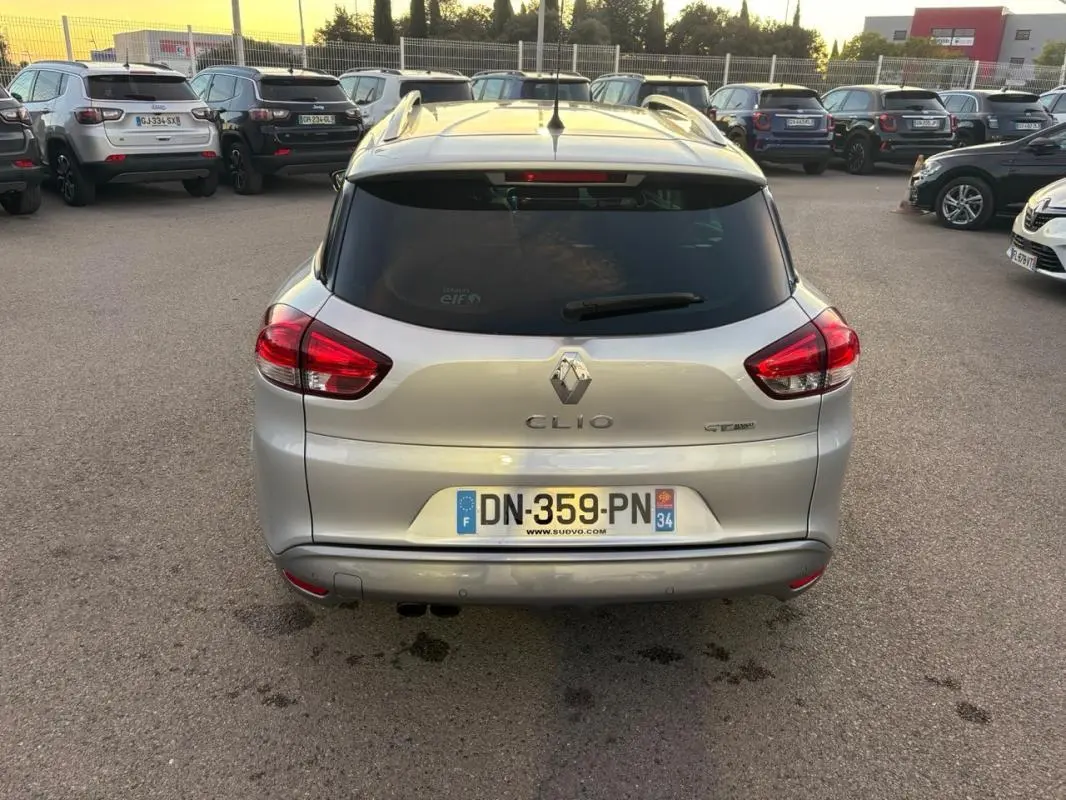 Vue arrière d'une Renault Clio IV Estate gris clair avec vitres surteintées et feux arrière allumés sur parking.