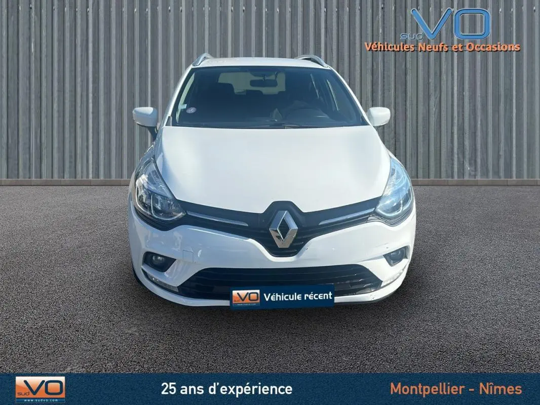 Vue frontale d'une Renault Clio IV Estate Business blanche avec phares allumés sur un sol goudronné.