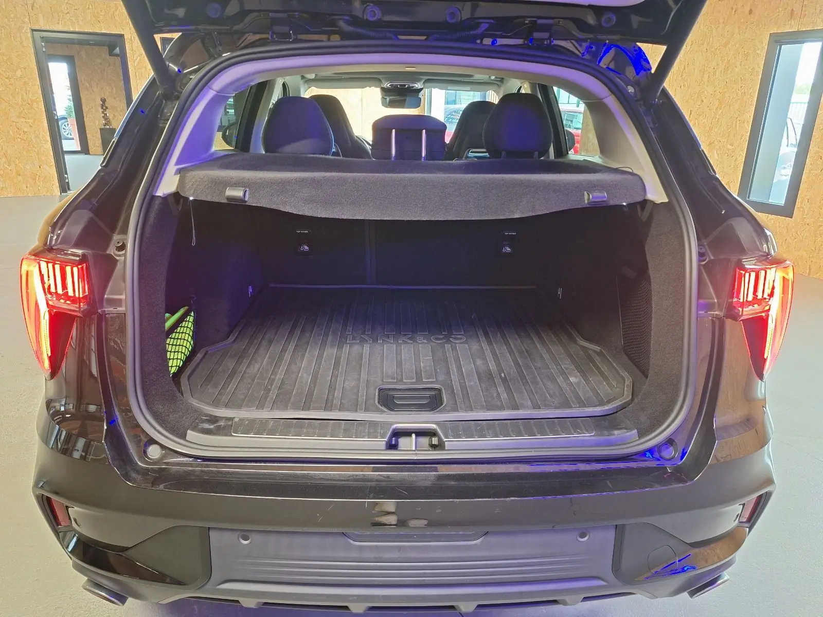 Vue arrière du coffre ouvert du LYNK & CO 01 noir avec tapis de coffre marqué et feux arrière allumés.