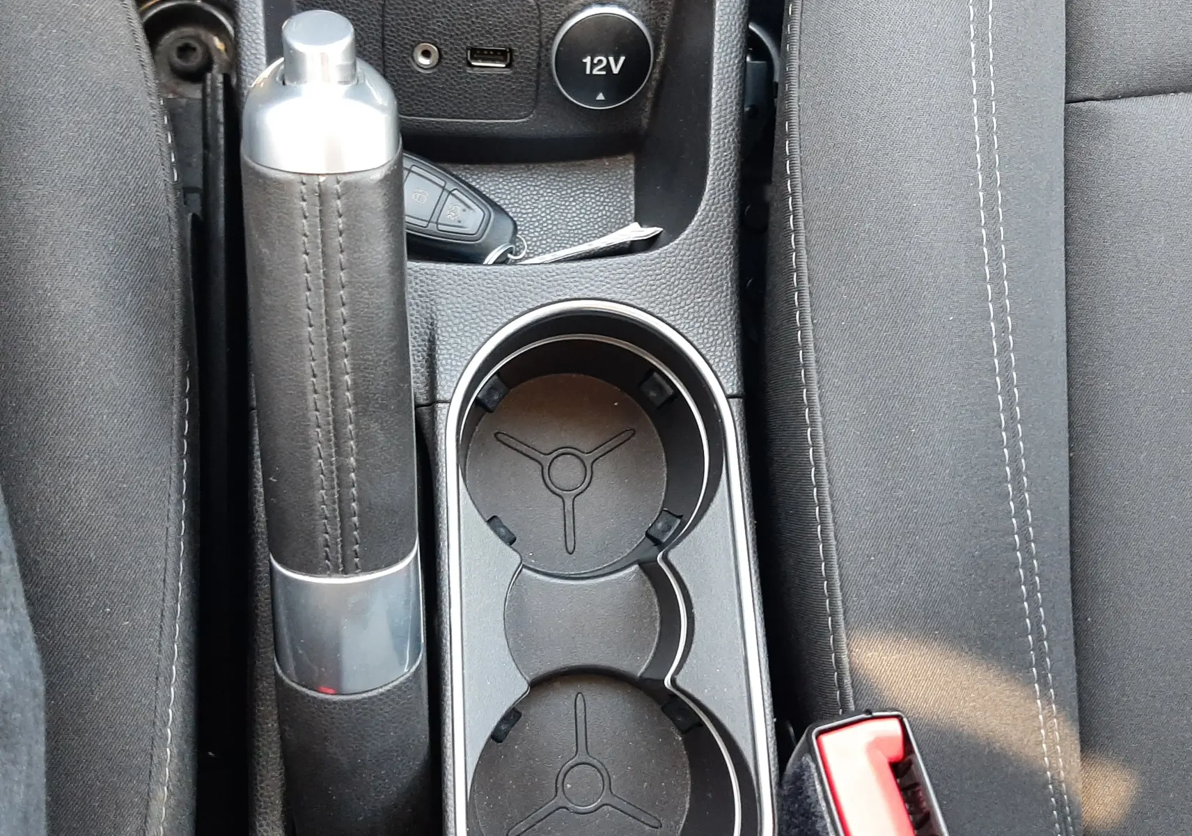 Vue plongeante sur la console centrale noire de la Ford Fiesta marron, avec frein à main, porte-gobelets et prise 12V visibles.