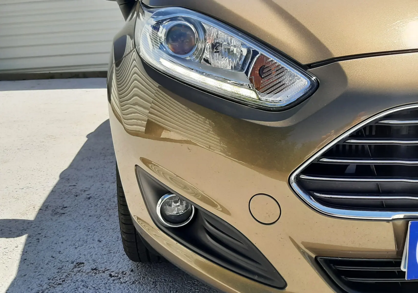Vue rapprochée avant gauche d'une Ford Fiesta marron métal, mettant en valeur le phare et la calandre chromée.