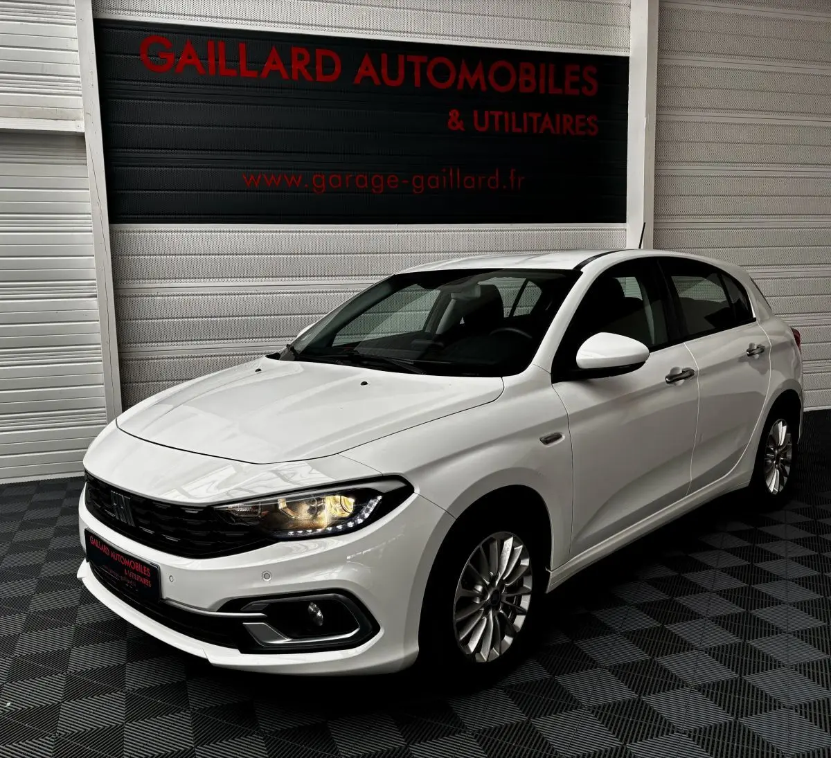 Vue 3/4 avant droite d'une Fiat Tipo blanche 2021 avec phares allumés dans un garage.