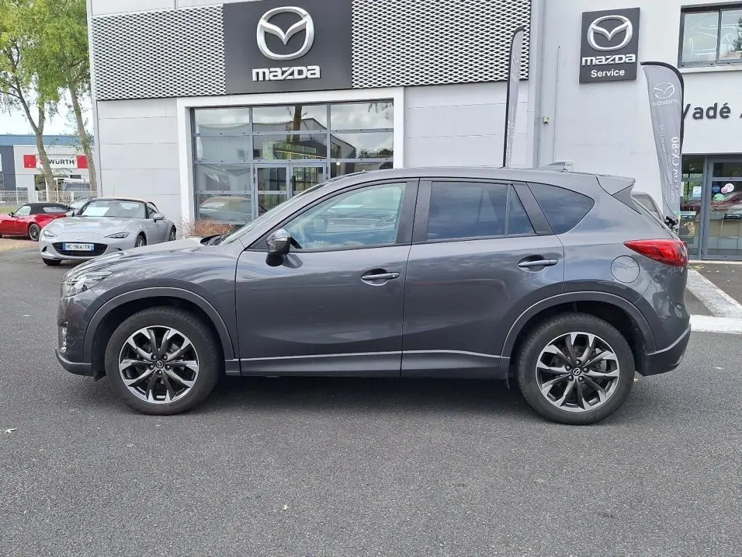 Profil côté gauche d'un Mazda CX-5 gris 2015 garé devant un concessionnaire Mazda avec jantes aluminium visibles.
