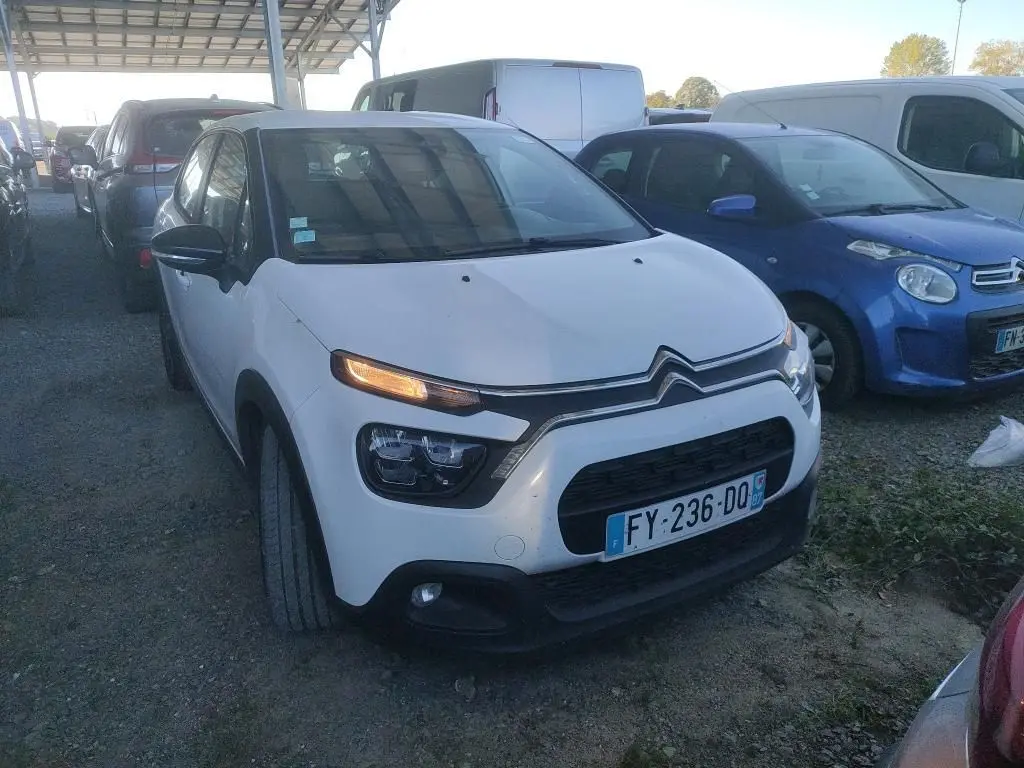 Vue 3/4 avant d'une Citroën C3 Société blanche de 2021 avec feux allumés, garée sur un sol en gravier.