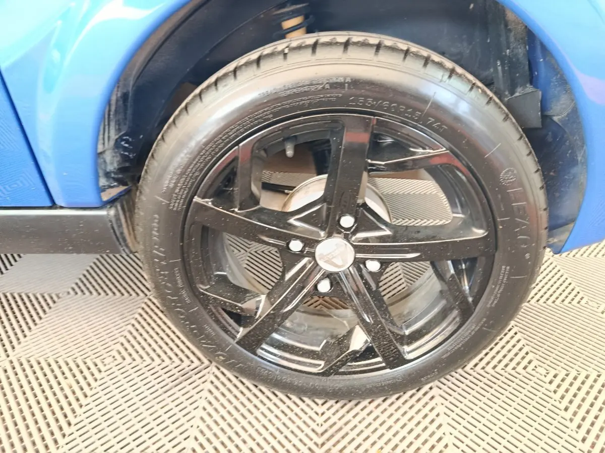 Gros plan sur la roue avant droite noire d'une Aixam Ambition 0.5 City Sport bleu 2024, avec partie de la carrosserie visible.