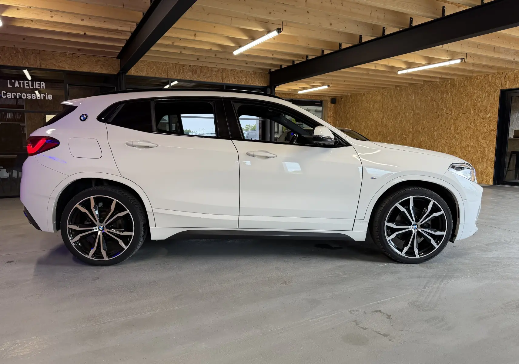 BMW X2 sDrive18d blanc vue de profil droit en intérieur, avec jantes alu 19'' et toit panoramique visible.