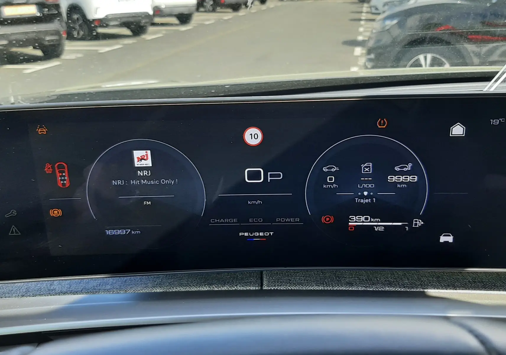 Vue rapprochée du tableau de bord numérique du Peugeot 5008 Hybrid 145ch, affichant la radio NRJ et les informations de conduite.