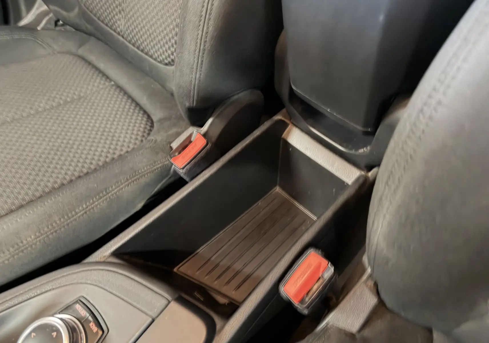 Vue rapprochée de la console centrale noire entre les sièges avant du BMW X1 F48 avec boutons de ceinture rouges visibles.