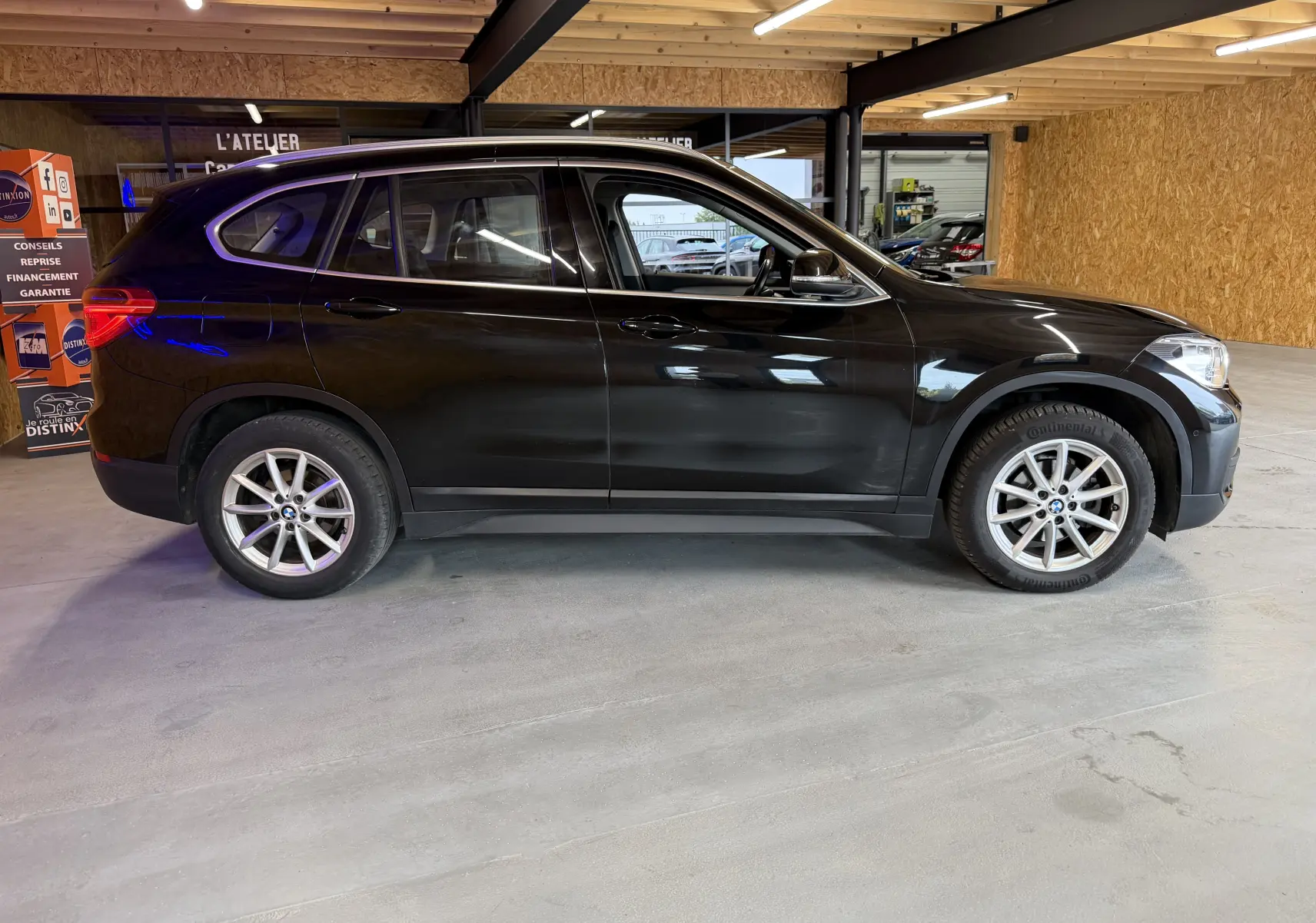 BMW X1 noir vue de profil côté gauche, stationné en intérieur avec jantes alliage et vitres teintées.