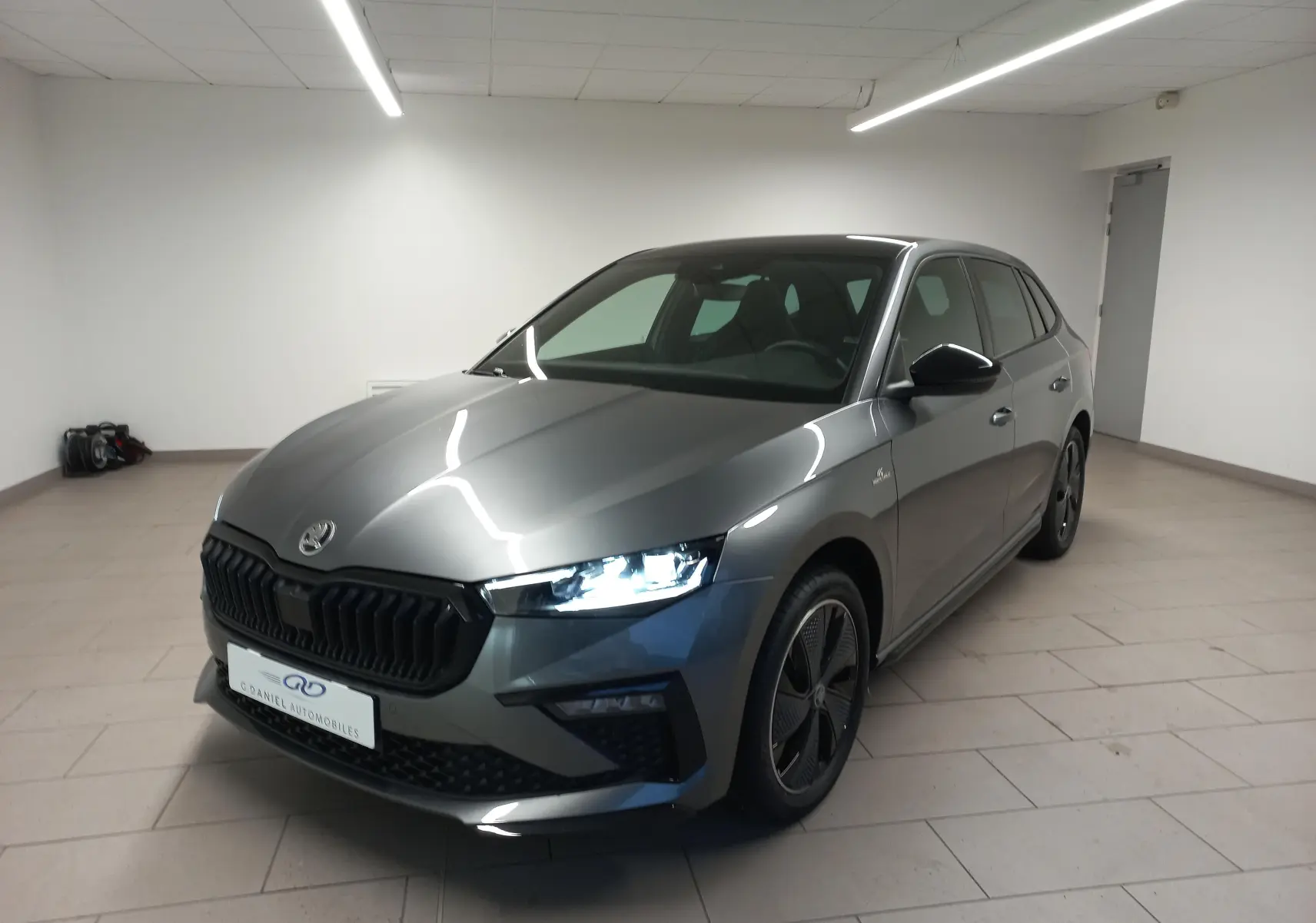 Vue 3/4 avant droite d'une Skoda Scala 1.5 TSI 150 DSG7 Monte-Carlo gris graphite aux phares allumés dans un garage.