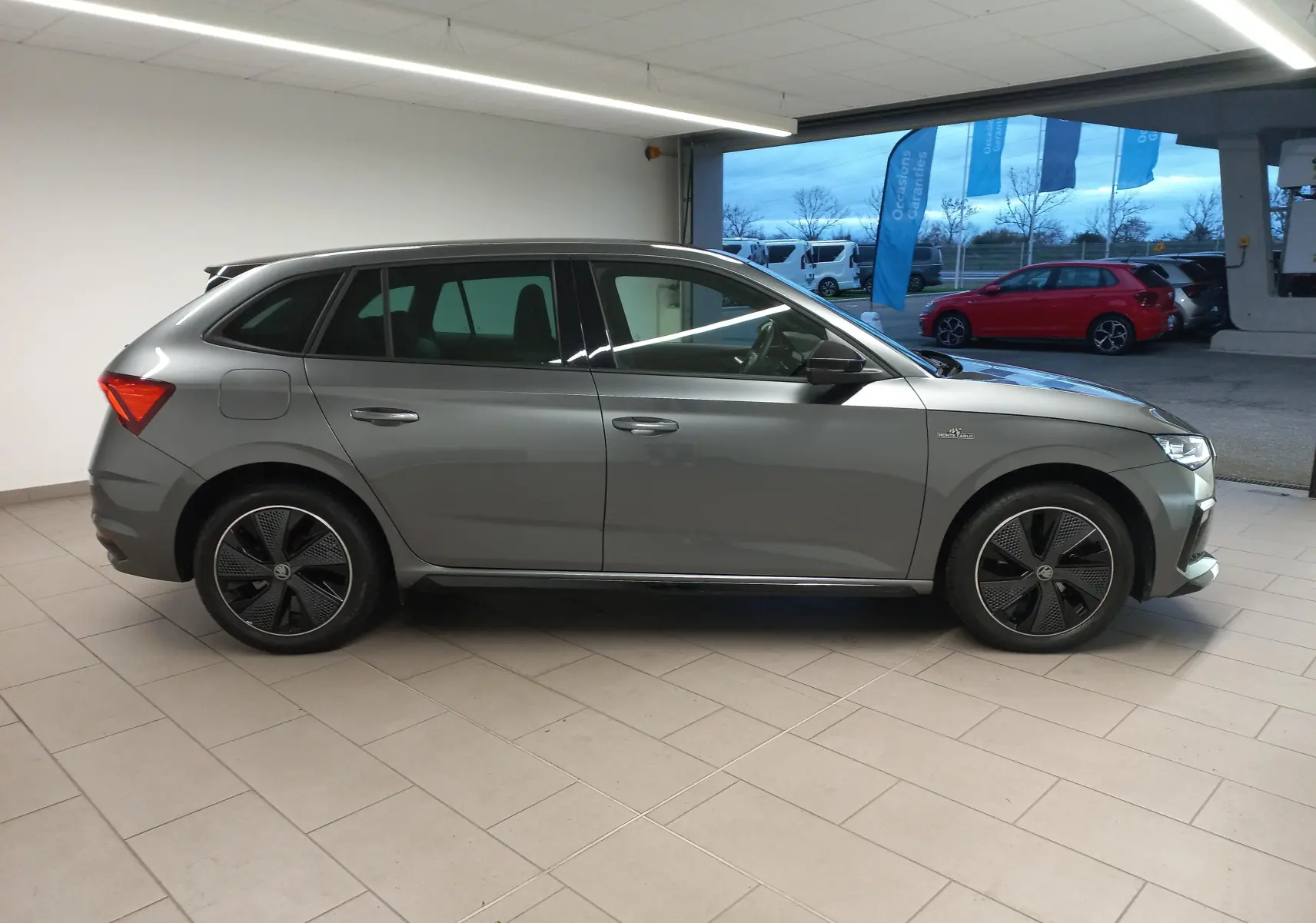 Profil côté gauche d'une Skoda Scala 2025 gris graphite avec jantes spécifiques Monte-Carlo en intérieur showroom.
