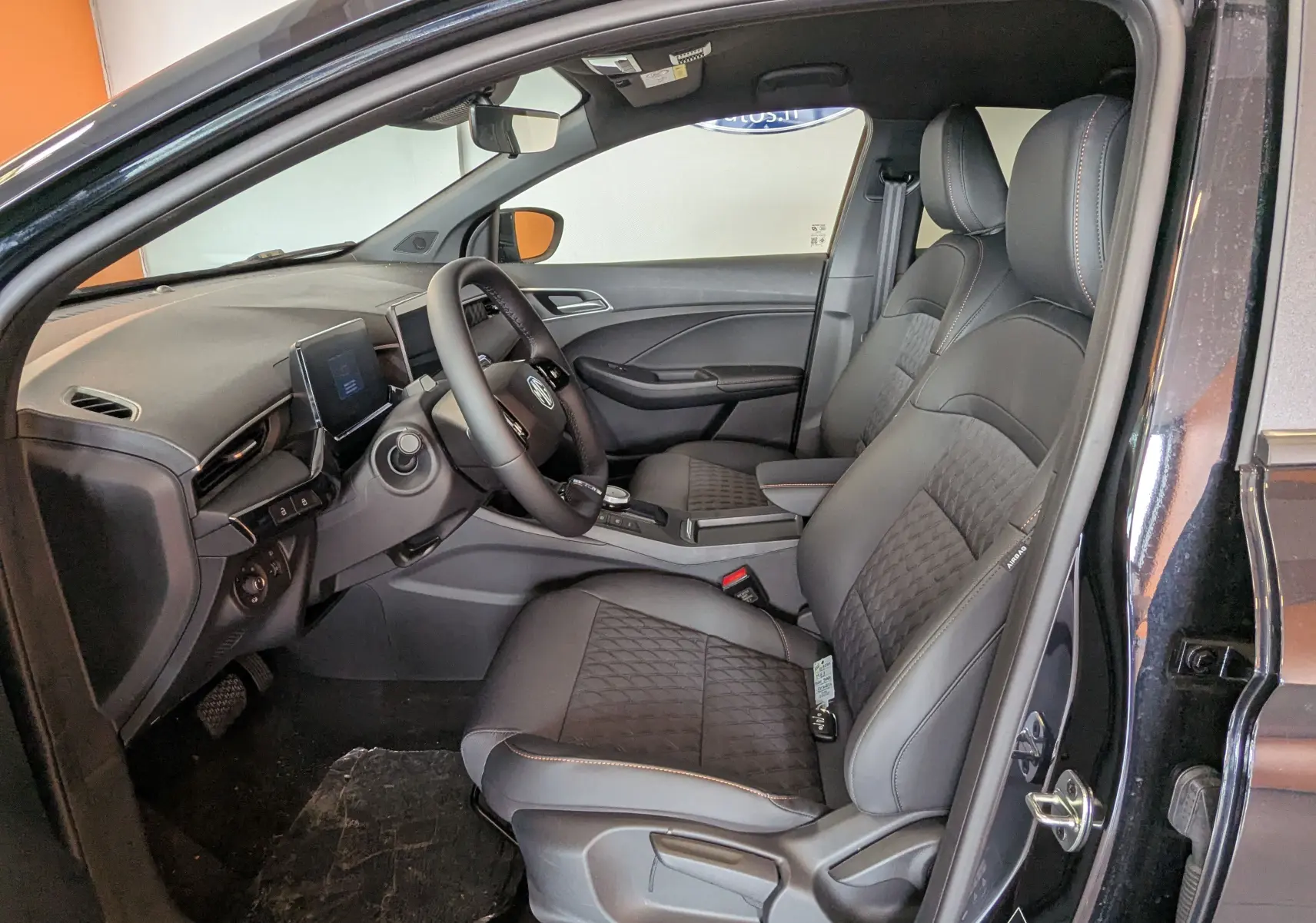 Vue intérieure côté conducteur de la MG3 1,5 HYBRID+ 2026 en Pebble Black, sièges noirs avec surpiqûres orange.