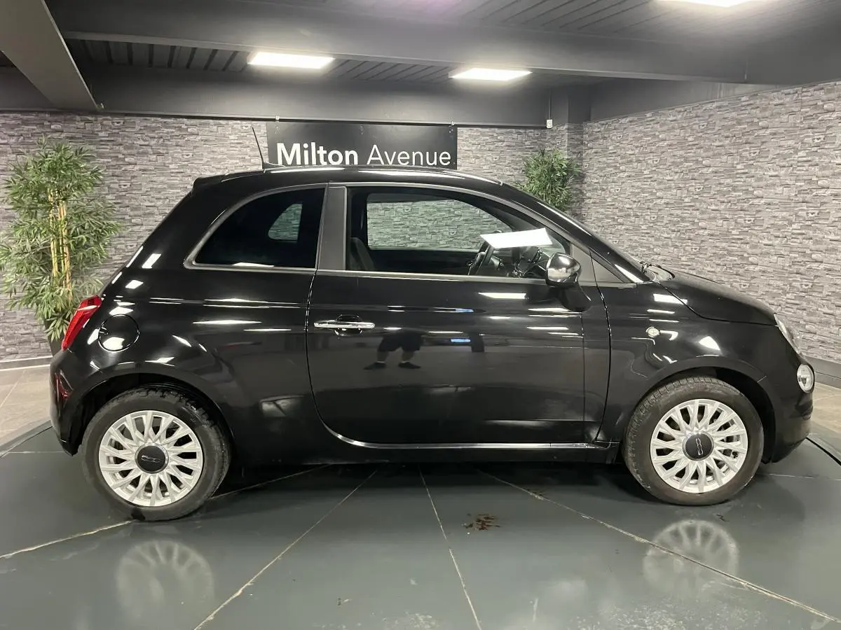 Profil droit de la Fiat 500 noire 2023 avec jantes alliage blanches et rétroviseur chromé en intérieur showroom.