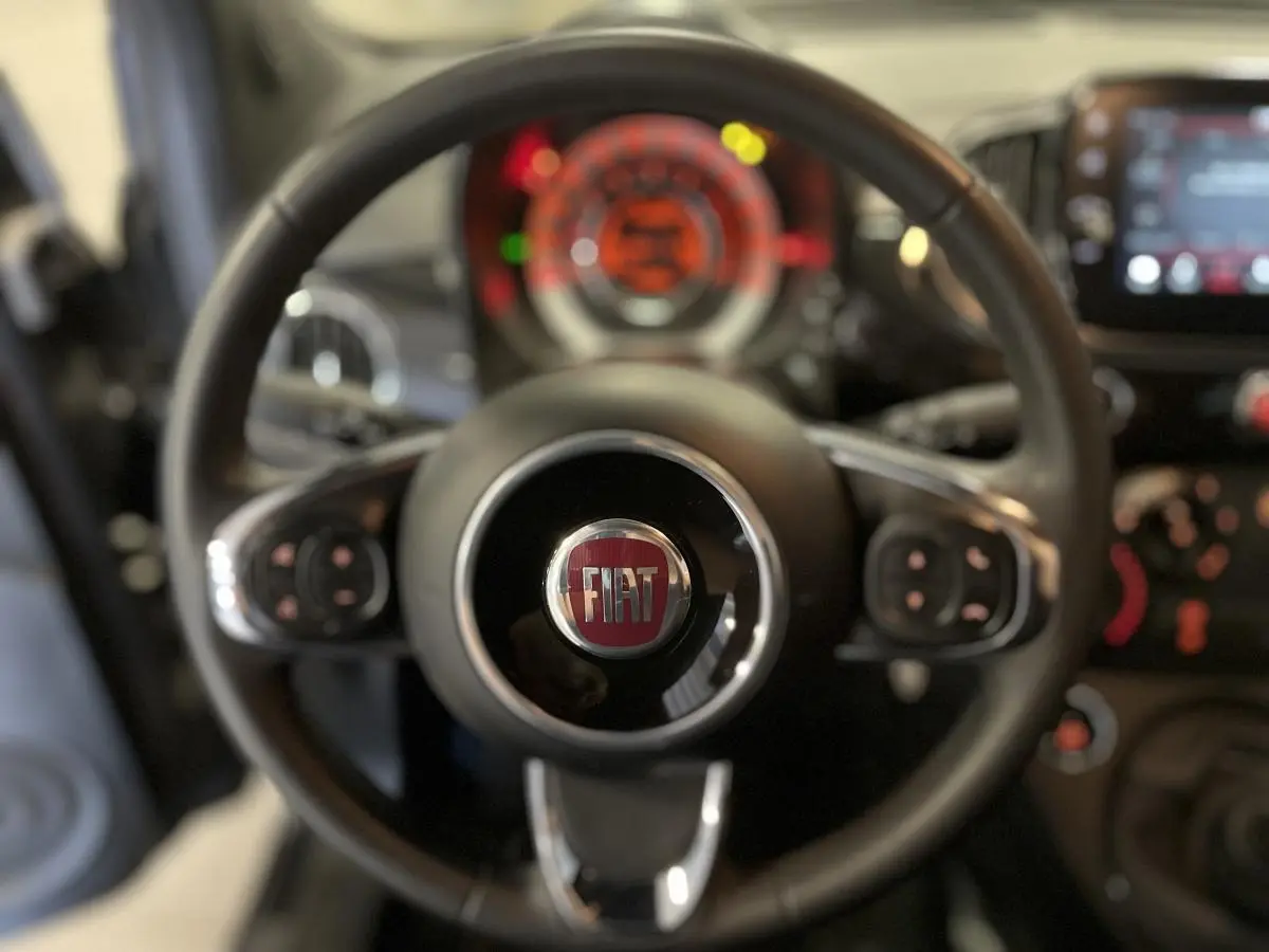 Volant noir de la Fiat 500 2023 avec logo central, commandes intégrées, tableau de bord flou en arrière-plan.