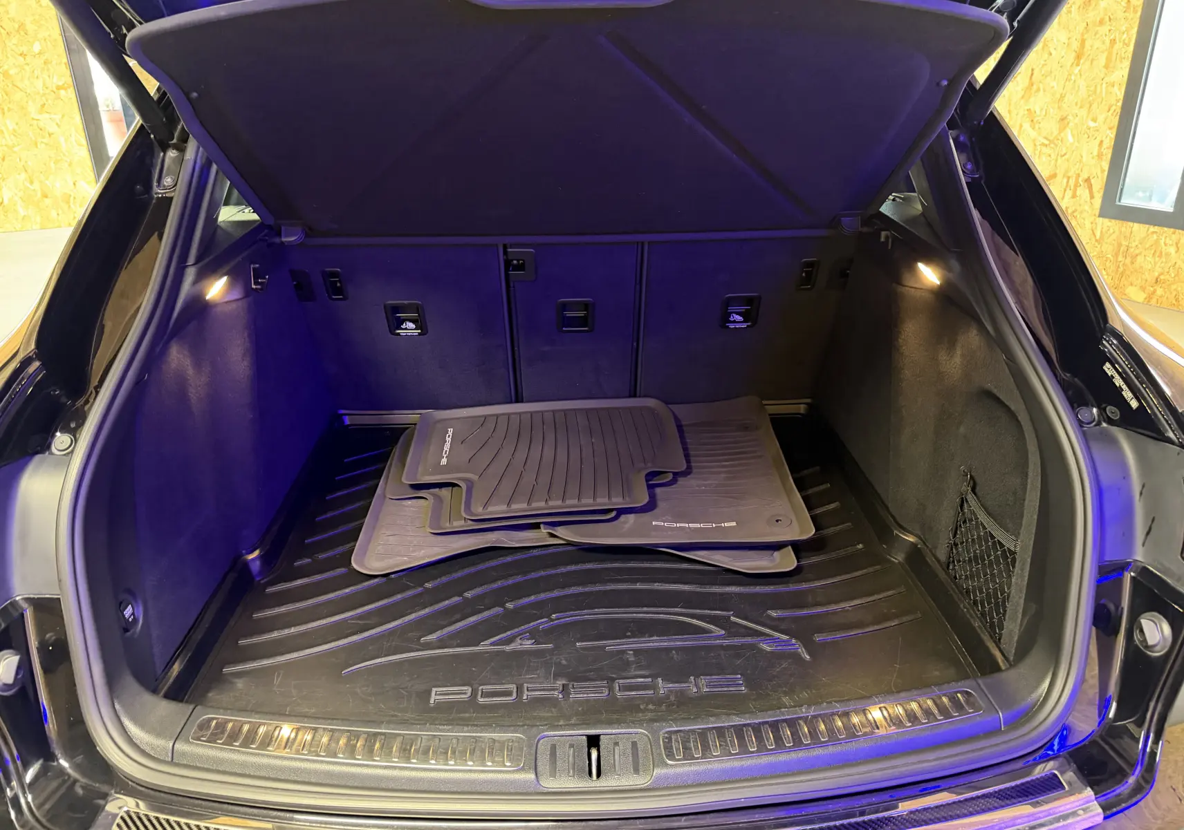 Coffre ouvert vu de l'arrière d'un Porsche Macan S noir avec tapis de sol Porsche et éclairage intérieur.