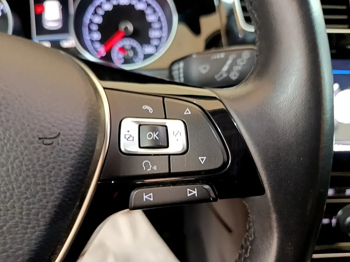 Gros plan sur les commandes gauche du volant cuir noir de la Volkswagen Golf gris clair, avec tableau de bord flou en arrière-plan.