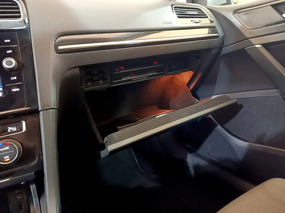 Gros plan sur la boîte à gants ouverte côté passager de la Volkswagen Golf gris clair, avec éclairage intérieur orange.