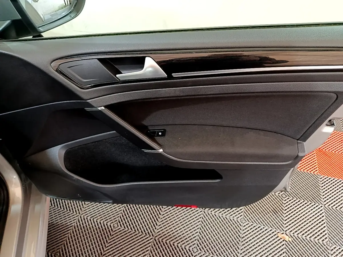 Vue rapprochée de la porte avant gauche d'une Volkswagen Golf gris clair, avec garniture noire et poignée argentée.