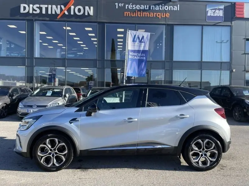 Renault Captur 1.6 E-Tech Hybrid 145 Intense gris métal vu de profil côté gauche sur parking devant concession multimarque.