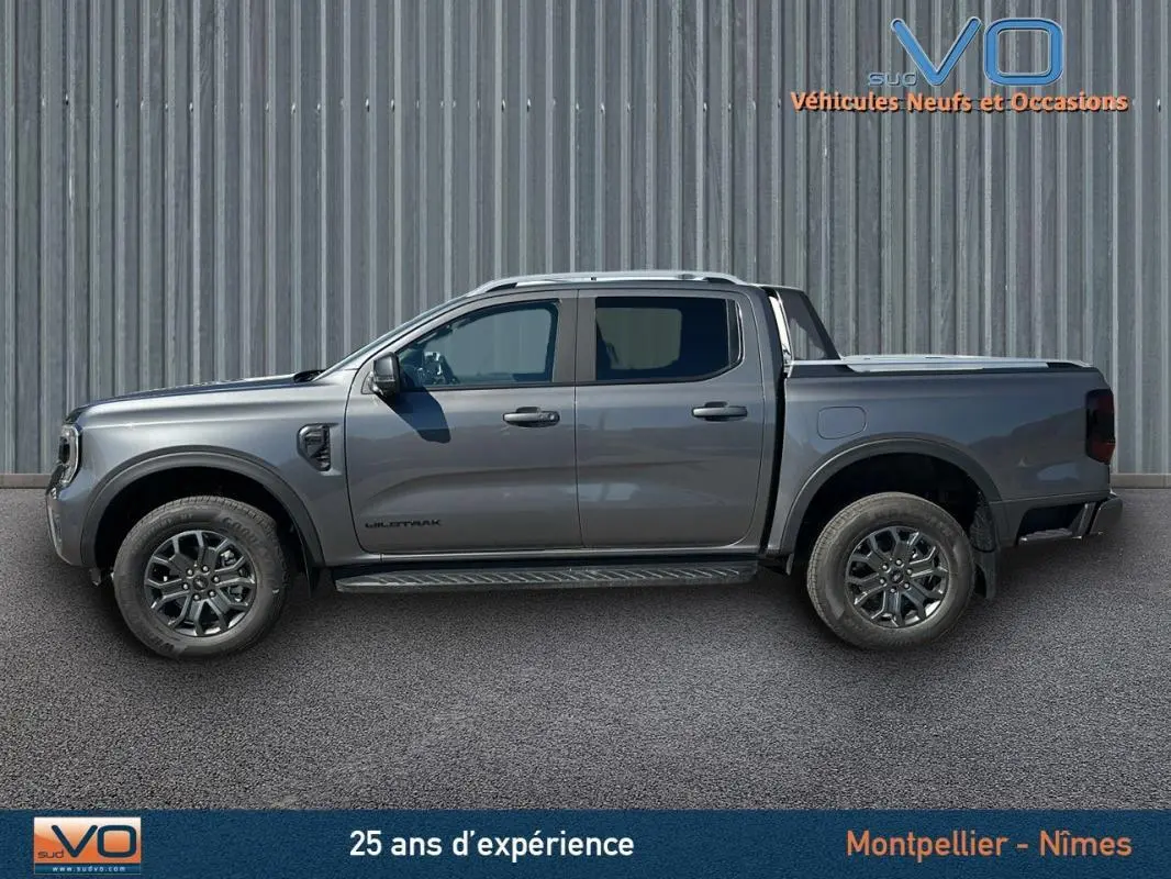 Profil côté gauche d'un Ford Ranger Wildtrak gris 2025 avec barres de toit et jantes alliage 17 pouces sur fond industriel.