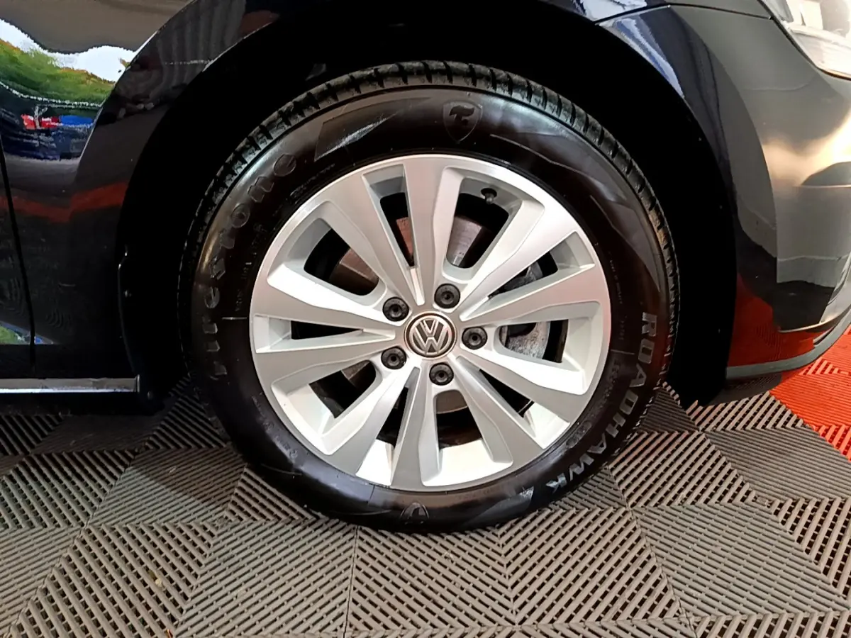 Gros plan sur la roue avant gauche d'une Volkswagen Golf noire, jante alliage argentée avec logo VW au centre.