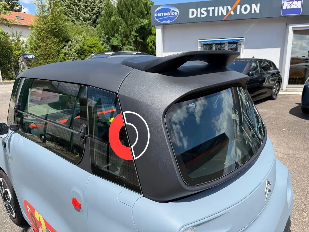 Vue 3/4 arrière droite d'une Citroën Ami MY AMI POP 2024 vert clair avec toit noir mat et logo rouge sur vitre arrière.