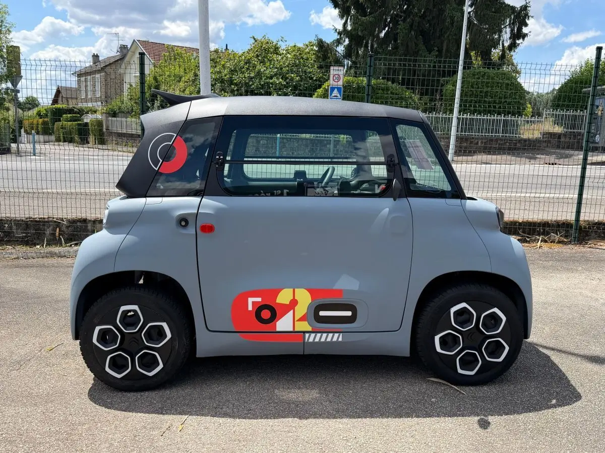 Vue de profil côté gauche d'une Citroën Ami MY AMI POP 2024 gris clair avec toit noir et décor graphique rouge et jaune sur la porte.