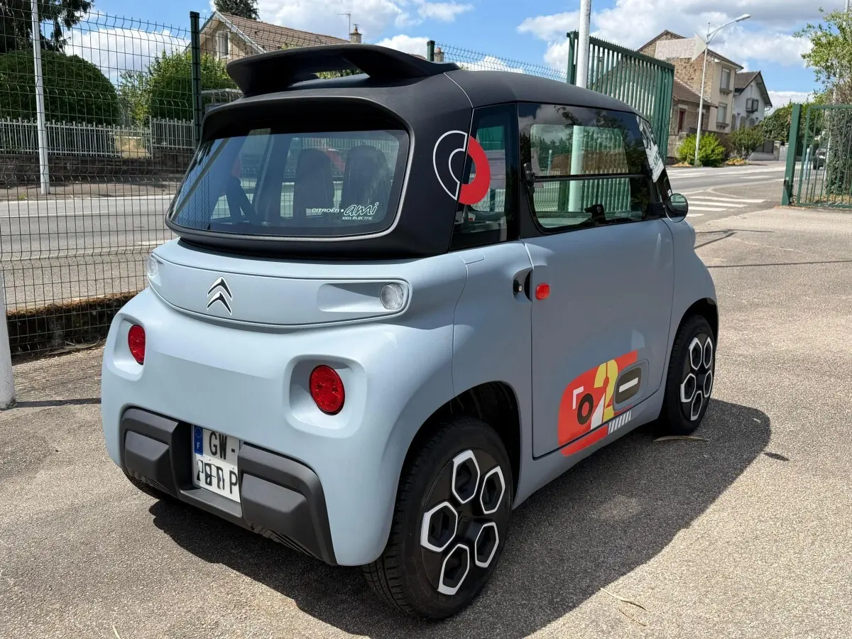 Vue 3/4 arrière droite d'une Citroën Ami MY AMI POP électrique gris clair avec toit noir et stickers colorés sur la porte.