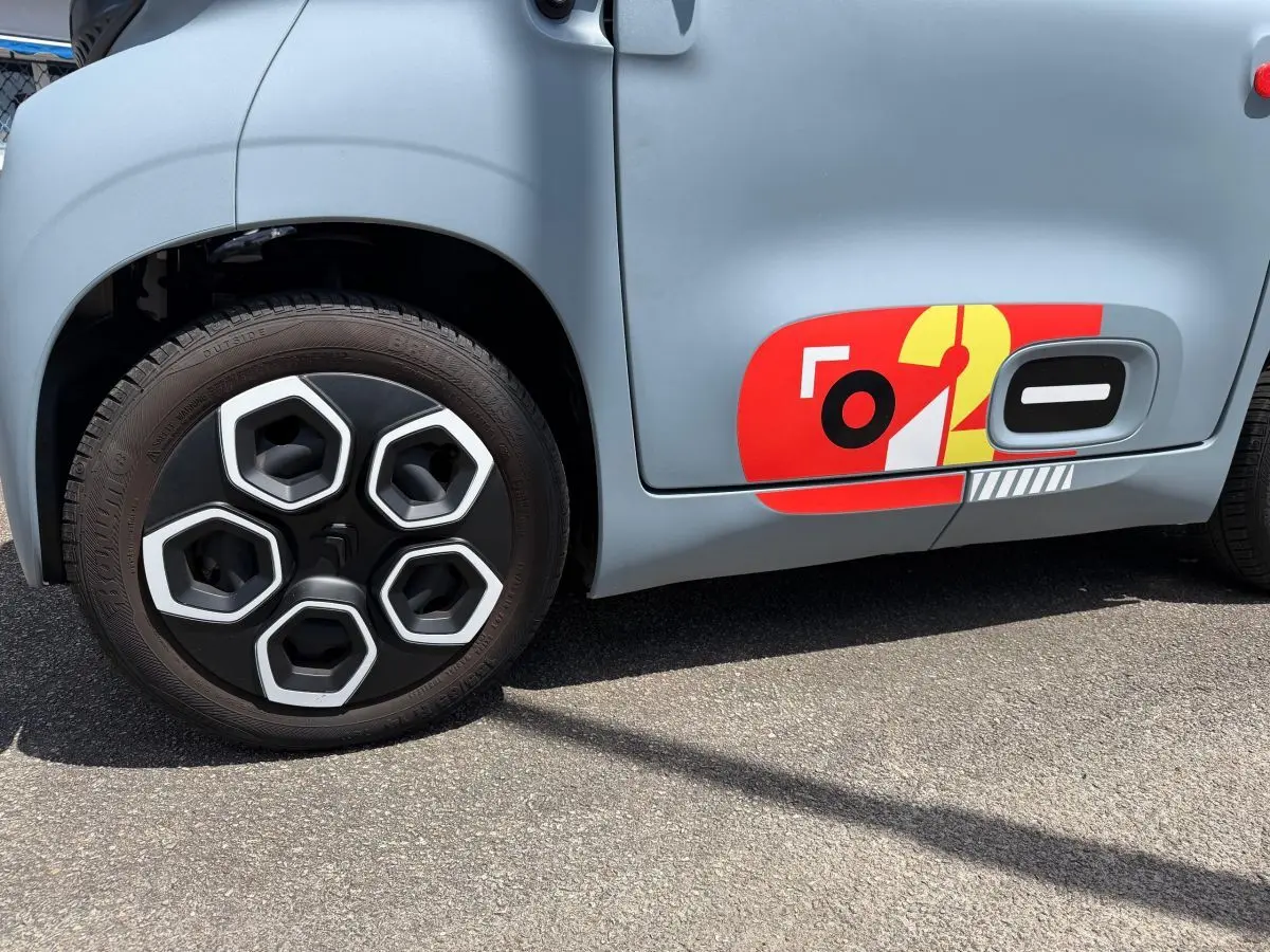 Gros plan sur la roue avant gauche et la porte côté conducteur d'une Citroën Ami MY AMI POP vert clair avec sticker rouge et jaune.