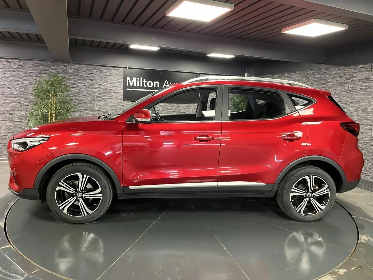 Vue de profil côté gauche du SUV MG ZS rouge métallisé avec jantes alliage noires et toit argenté en intérieur showroom.