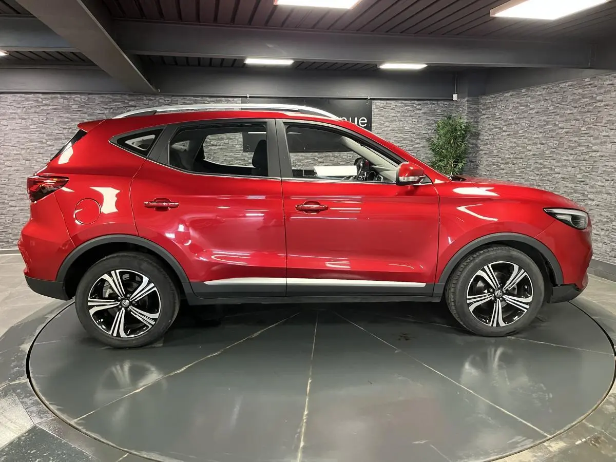Vue latérale droite du SUV MG ZS rouge métallisé 2022 avec jantes alliage noires et toit argenté dans un showroom.
