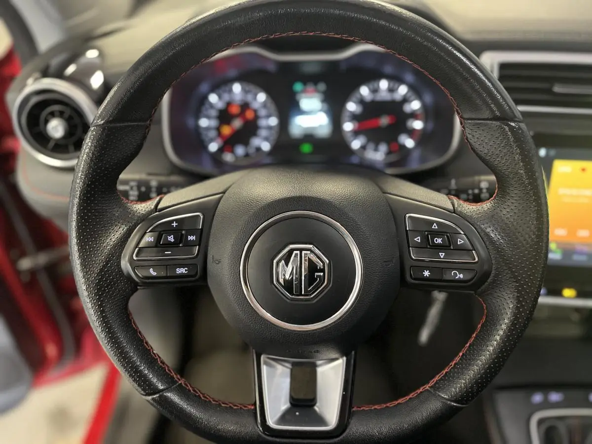 Gros plan sur le volant cuir noir avec surpiqûres rouges du MG ZS rouge, tableau de bord et commandes visibles en arrière-plan.