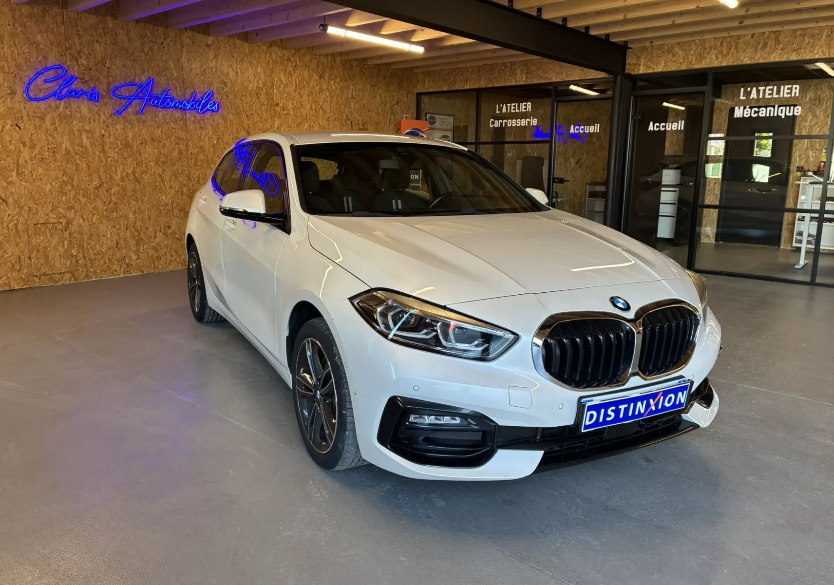 BMW Série 1 118i Edition Sport blanche vue en 3/4 avant droit dans un showroom avec calandre noire distinctive.