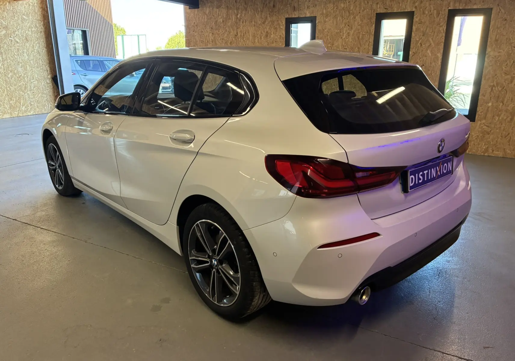 BMW Série 1 118i Edition Sport blanche vue en 3/4 arrière droit dans un garage avec jantes bi-ton et feux LED.