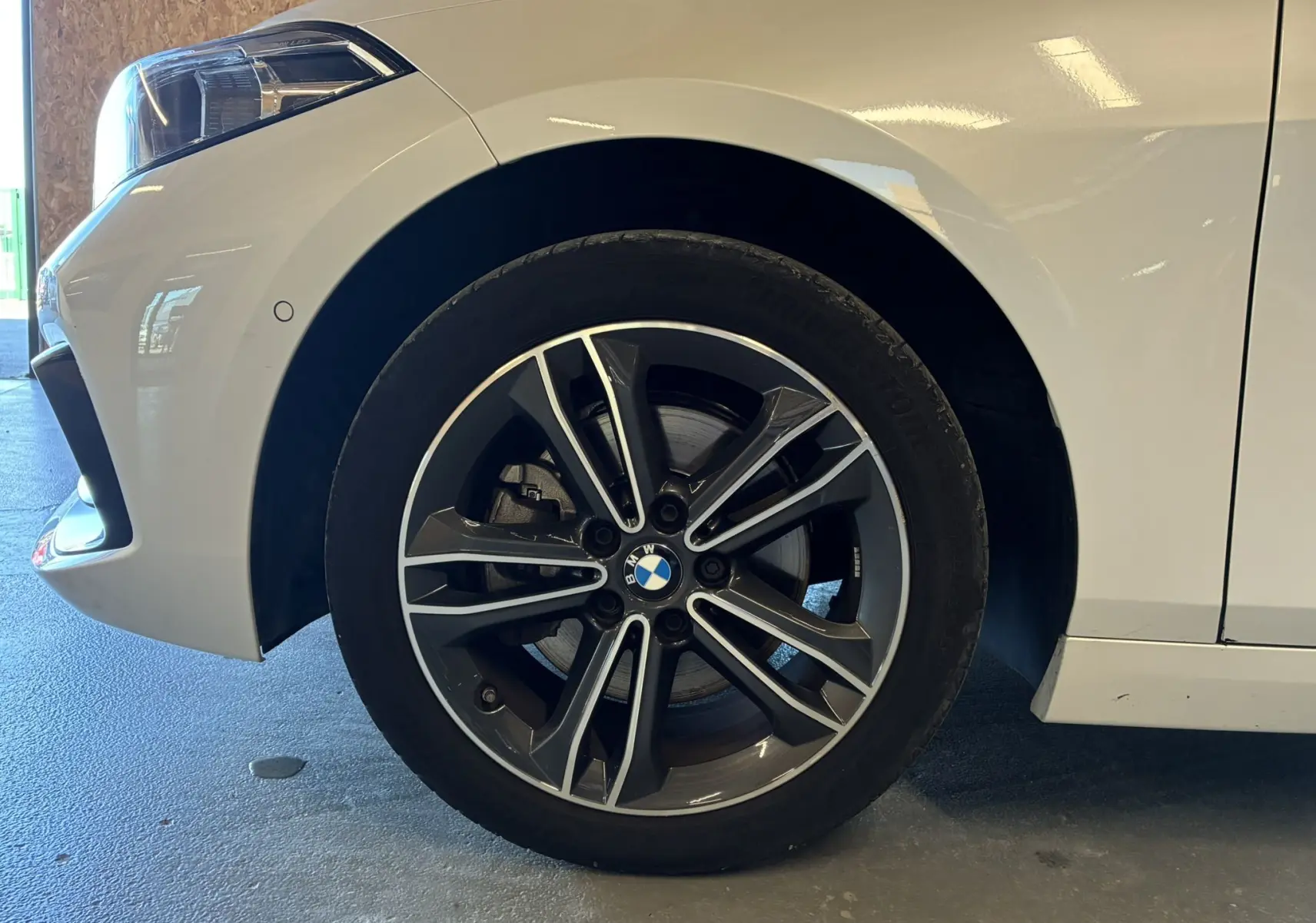 Gros plan sur la roue avant gauche d'une BMW Série 1 blanche avec jante alliage bicolore et phare avant partiel visible.