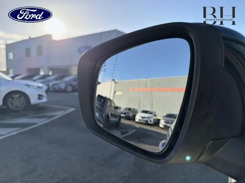 Rétroviseur côté droit montrant le reflet d'un parking avec plusieurs véhicules, prise en extérieur au soleil.