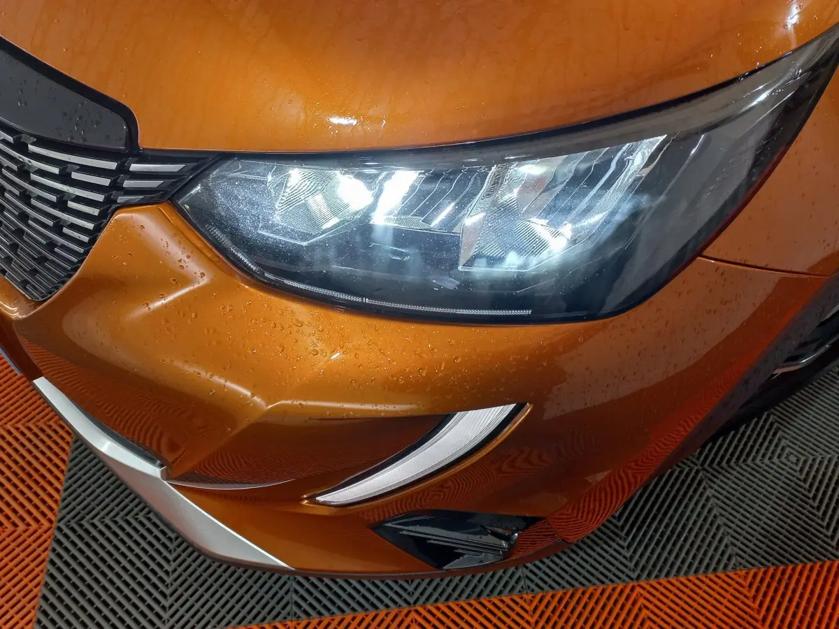 Gros plan sur le phare avant droit et la calandre d’une Peugeot 2008 orange métallisé avec gouttes d’eau visibles.