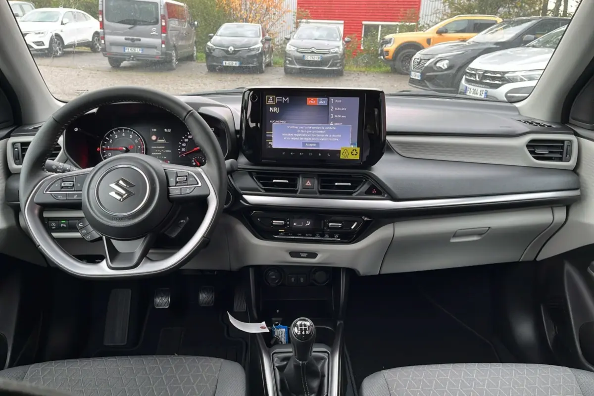 Vue intérieure avant du Suzuki Swift 2026 gris, volant cuir multifonctions et écran tactile central avec boîte manuelle.